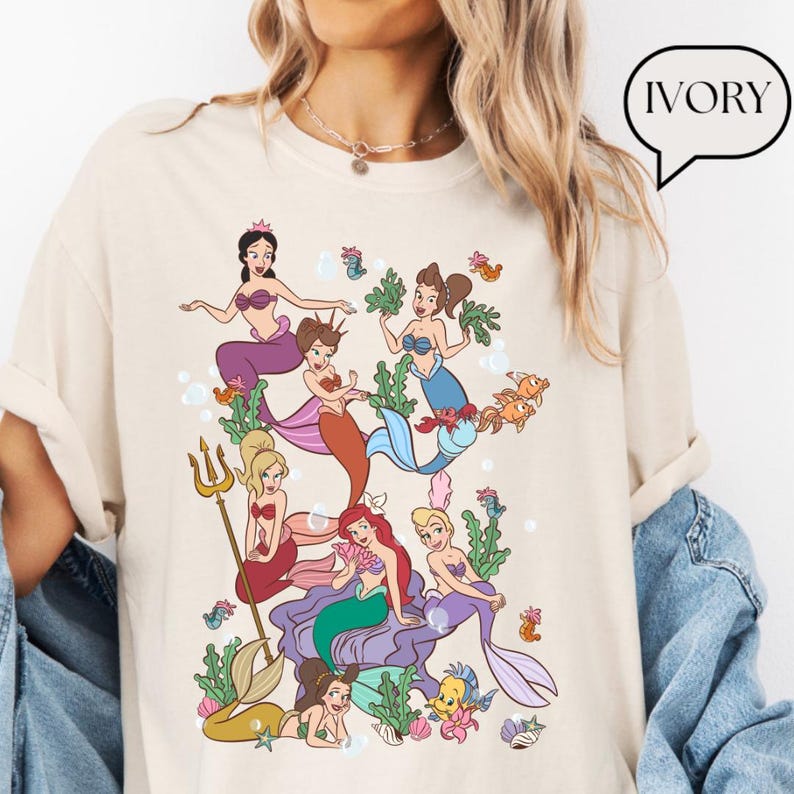 Op de afbeelding: Ivoor kleurig t-shirt met een kleurrijke illustratie van zeemeerminnen. De afbeelding toont verschillende zeemeerminnen met verschillende haar- en staartkleuren, omringd door waterplanten, bellen en vissen. Het woord "IVORY" staat in een tekstballon.