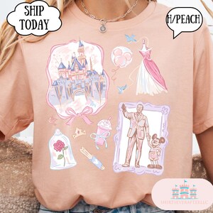 Op de afbeelding: Perzikkleurig T-shirt met diverse illustraties, waaronder een kasteel, jurk, ballonnen en een ingelijste afbeelding van een man met Mickey Mouse. Het shirt heeft ook de tekst "SHIP TODAY" en "H/PEACH".