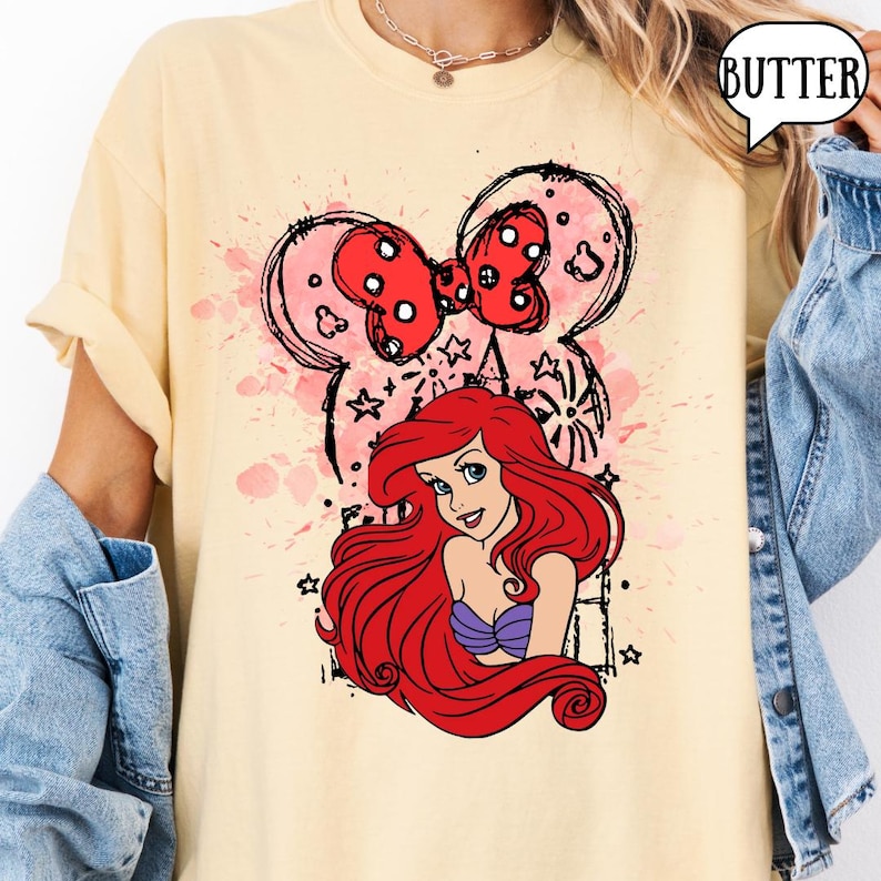 May include: Cream-colored t-shirt featuring a graphic of Ariel from The Little Mermaid, with a red bow and Mickey Mouse ears design. The shirt has a distressed, vintage look with pink splatter details and black outlines. The word "BUTTER" is in a speech bubble.
