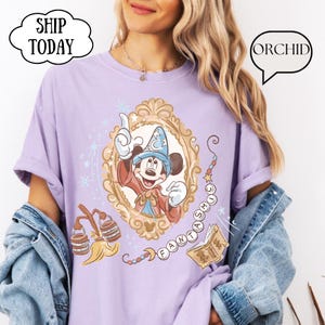 K&ouml;nnte beinhalten: Ein helllila T-Shirt mit einer gerahmten Illustration von Mickey Mouse in einem Zauberer-Outfit. Das Design enth&auml;lt das Wort "FANTASMIC" und andere magische Elemente. Das Shirt hat auch die W&ouml;rter "ORCHID" und "SHIP TODAY" in Sprechblasen.