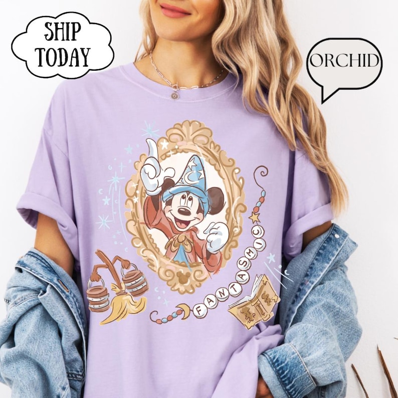 K&ouml;nnte beinhalten: Ein helllila T-Shirt mit einer gerahmten Illustration von Mickey Mouse in einem Zauberer-Outfit. Das Design enth&auml;lt das Wort "FANTASMIC" und andere magische Elemente. Das Shirt hat auch die W&ouml;rter "ORCHID" und "SHIP TODAY" in Sprechblasen.