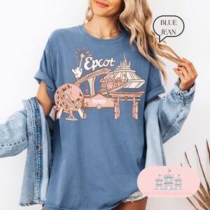 May include: A blue jean t-shirt with a pink and white graphic of Epcot, featuring the Spaceship Earth ride, the World Showcase, and the Frozen Ever After ride. The text "Epcot" is written in a pink font.