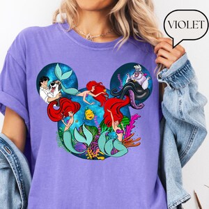 Puede incluir: Una camiseta violeta con un dise&ntilde;o gr&aacute;fico de los personajes de La Sirenita de Disney. El dise&ntilde;o incluye a Ariel, &Uacute;rsula y otros personajes dentro de una forma de oreja de Mickey Mouse. La palabra "VIOLET" est&aacute; en una burbuja.