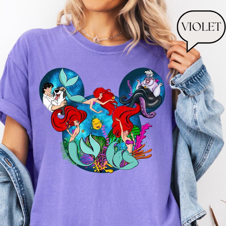 Puede incluir: Una camiseta violeta con un dise&ntilde;o gr&aacute;fico de los personajes de La Sirenita de Disney. El dise&ntilde;o incluye a Ariel, &Uacute;rsula y otros personajes dentro de una forma de oreja de Mickey Mouse. La palabra "VIOLET" est&aacute; en una burbuja.