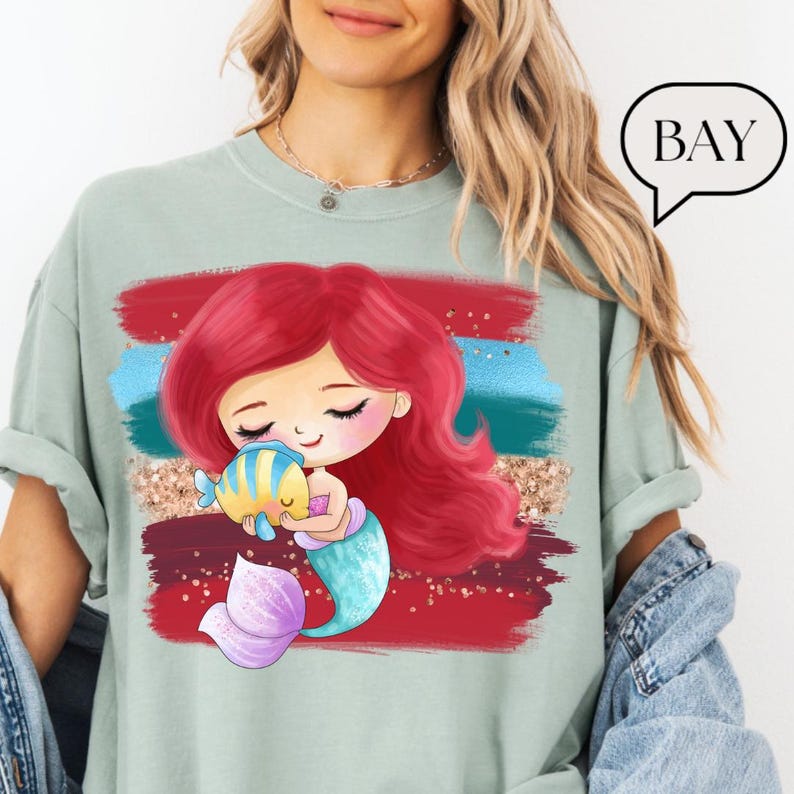 Puede incluir: Camiseta verde claro con un dise&ntilde;o de sirena de dibujos animados. La sirena tiene el pelo rojo, una cola azul y morada, y sostiene un pez amarillo y azul. El dise&ntilde;o incluye pinceladas rojas, azules y doradas. La palabra "BAY" est&aacute; en un bocadillo.