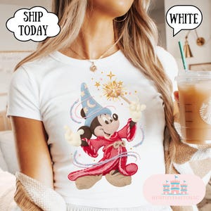 May include: White t-shirt with a colourful illustration of Mickey Mouse as a wizard, holding a magic wand. The design includes a blue wizard hat with a crescent moon, a red robe, and swirling lines. The text "SHIP TODAY" is in a speech bubble.