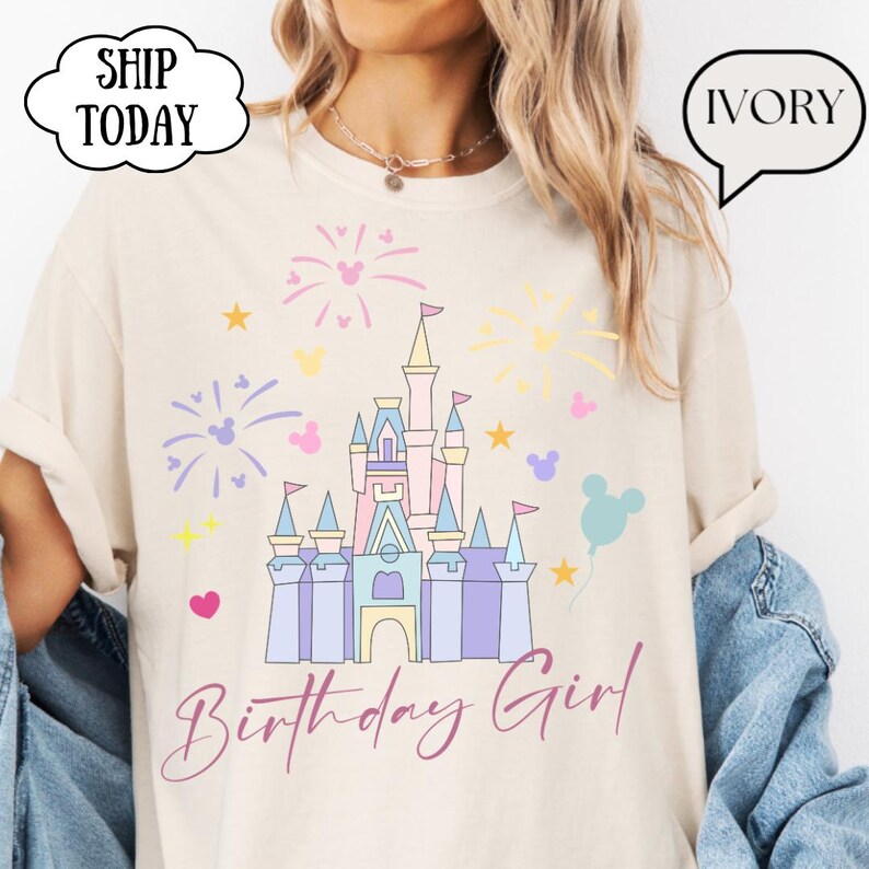 May include: Ivory t-shirt with a pastel castle graphic, fireworks, stars, and Mickey Mouse silhouettes. The text "Birthday Girl" is written in a cursive font. The shirt has rolled-up sleeves.