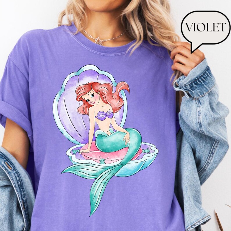 May include: A violet t-shirt with a cartoon mermaid design. The mermaid has red hair, a purple seashell bra, and a teal tail, seated in an open seashell on a pink pillow. The word "VIOLET" is in a speech bubble.