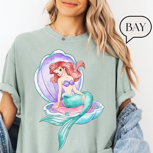 May include: A pale green t-shirt with a watercolour illustration of a mermaid with red hair, a purple seashell, and the word "BAY" in a speech bubble. The mermaid has a teal tail and a purple seashell bra.