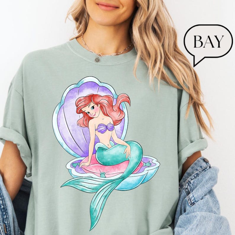 May include: A pale green t-shirt with a watercolour illustration of a mermaid with red hair, a purple seashell, and the word "BAY" in a speech bubble. The mermaid has a teal tail and a purple seashell bra.