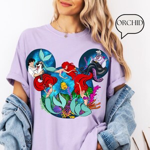 Puede incluir: Una camiseta de color lila claro con un gr&aacute;fico colorido de personajes de La Sirenita. El dise&ntilde;o est&aacute; dispuesto dentro de la forma de las orejas de Mickey Mouse, con Ariel, el Pr&iacute;ncipe Eric, &Uacute;rsula y otros personajes. La palabra "ORCHID" est&aacute; en una burbuja.
