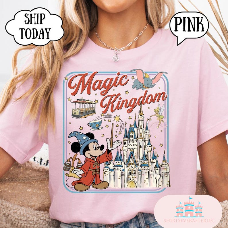 May include: Pink t-shirt featuring a graphic of Mickey Mouse as a wizard with a castle, Dumbo, and the words "Magic Kingdom." The design is framed with a light blue border and stars. The shirt has a round neckline.