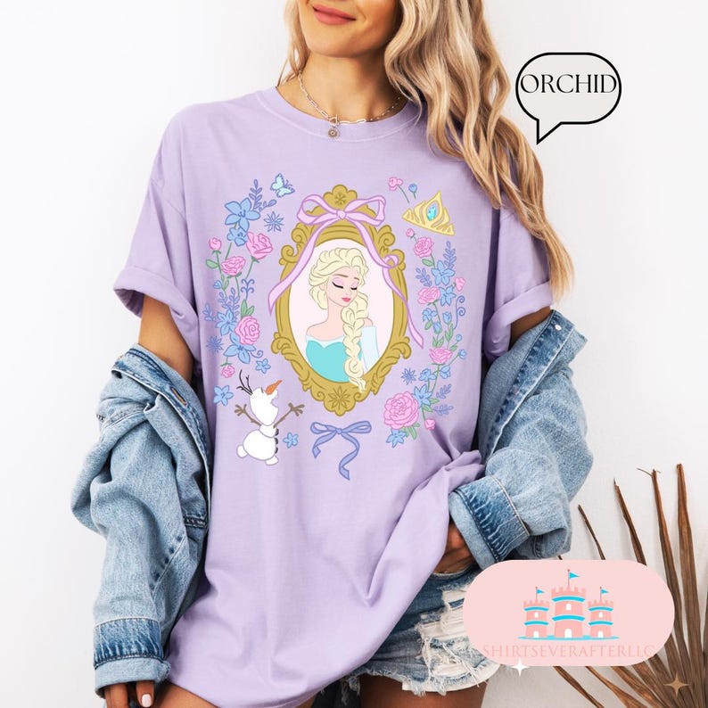 K&ouml;nnte beinhalten: Lilafarbenes T-Shirt mit einem Blumendesign, das ein gerahmtes Bild von Elsa aus Frozen, eine Krone und Olaf zeigt. Der Text "ORCHID" befindet sich in einer Sprechblase.