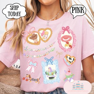Op de afbeelding: Zachtroze T-shirt met illustraties van stripfiguren in sierlijke kaders. Het shirt bevat de zin "TO INFINITY AND BEYOND" en het woord "PINK" in een tekstballon. Het shirt heeft ook de tekst "SHIP TODAY" in een tekstballon.