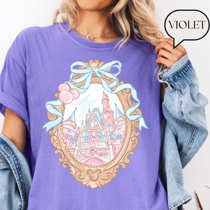 Puede incluir: Camiseta violeta con un dise&ntilde;o de castillo enmarcado, cinta azul claro y detalles en rosa. El marco tiene un borde dorado con una cabeza de Mickey Mouse en la parte inferior. La palabra "VIOLET" est&aacute; en un bocadillo.
