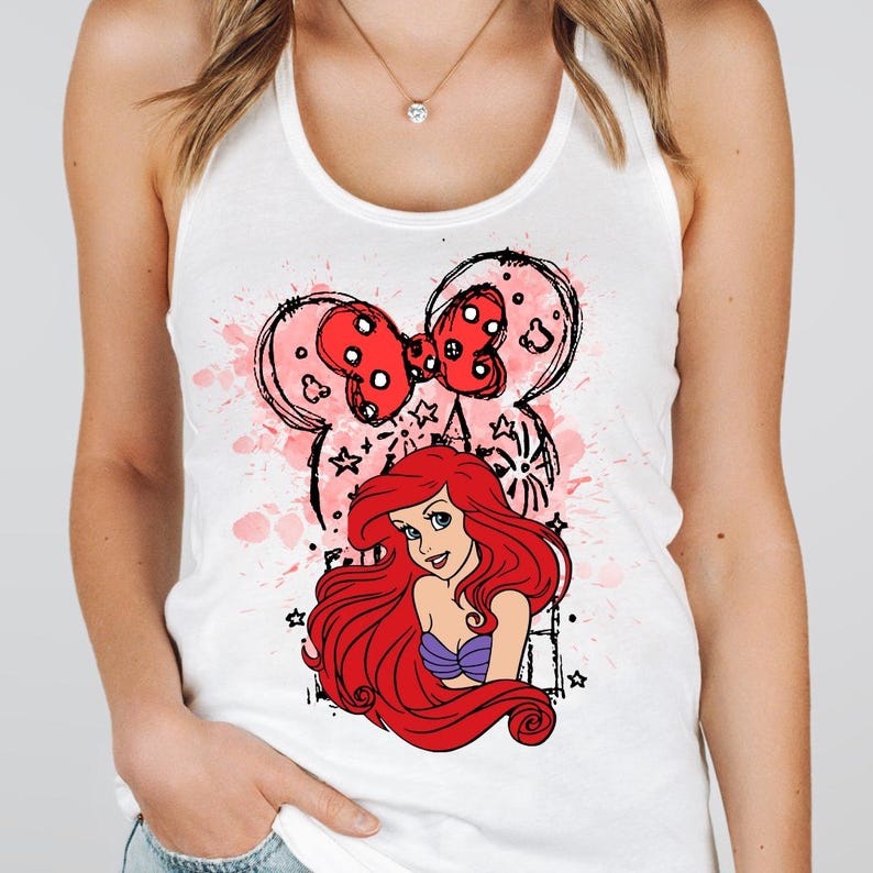 May include: White tank top featuring a graphic of Ariel from The Little Mermaid. The design includes a red bow, Minnie Mouse ears, and a pink splatter effect. Ariel has long red hair and a purple top.
