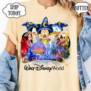 May include: Butter-colored t-shirt featuring a colorful graphic of Mickey Mouse, Goofy, Donald Duck, and Pluto in front of a castle and other Disney World landmarks. The words "Walt Disney World" are printed below the graphic.