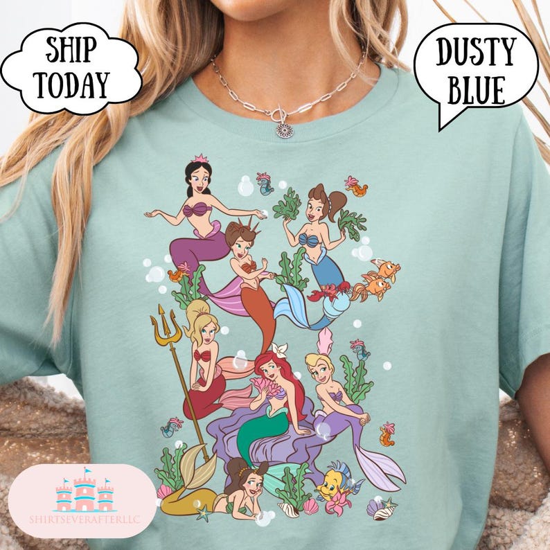 Op de afbeelding: Lichtblauw T-shirt met een kleurrijke illustratie van zeemeerminnen. De afbeelding toont verschillende zeemeerminnen met diverse haar- en kledingstijlen, omringd door waterplanten en vissen. Het shirt heeft ook de tekst "Dusty Blue" en "Ship Today".