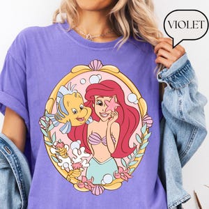 May include: Violet-colored t-shirt featuring a cartoon illustration of Ariel from The Little Mermaid, holding a starfish with Flounder. The graphic is framed in a gold-colored oval with pink and green accents. The word "VIOLET" is in a speech bubble.