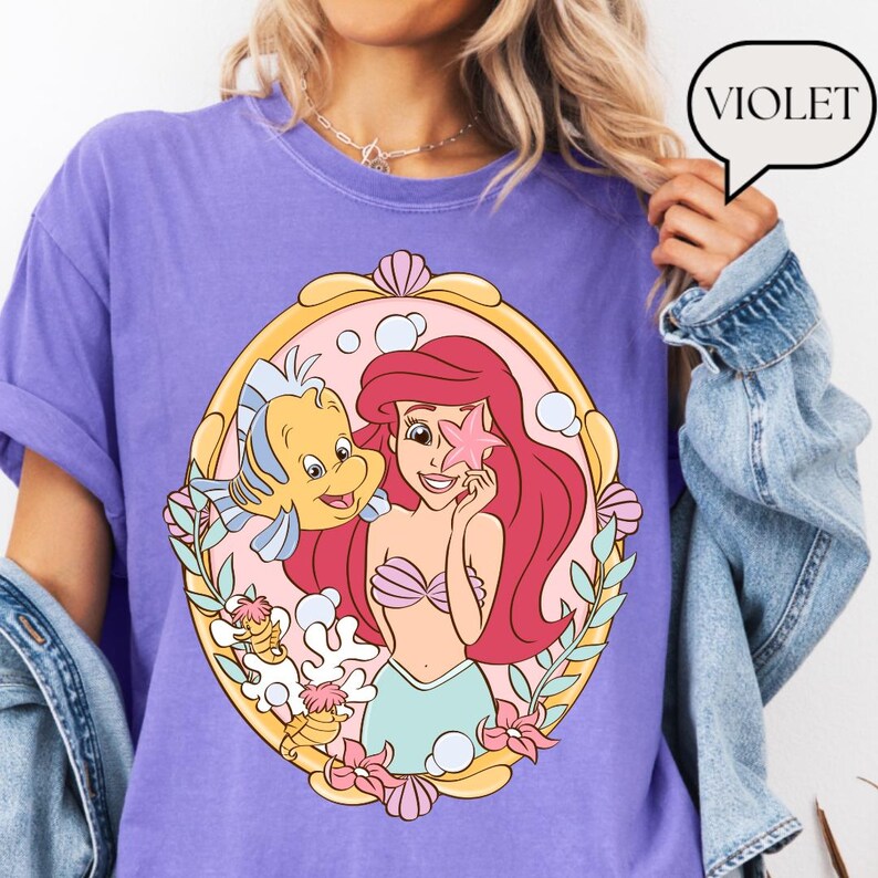 May include: Violet-colored t-shirt featuring a cartoon illustration of Ariel from The Little Mermaid, holding a starfish with Flounder. The graphic is framed in a gold-colored oval with pink and green accents. The word "VIOLET" is in a speech bubble.