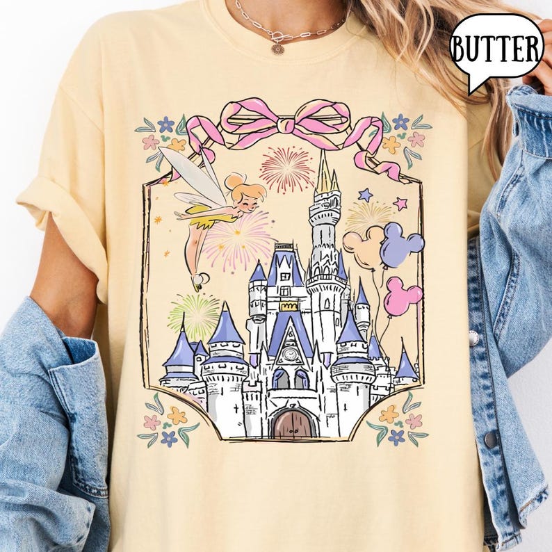 Puede incluir: Camiseta amarilla clara con una ilustraci&oacute;n de un castillo de cuento de hadas. El dise&ntilde;o incluye torres azules, fuegos artificiales y Campanilla. Tambi&eacute;n se presenta un lazo rosa y globos. La palabra "BUTTER" est&aacute; en una burbuja.