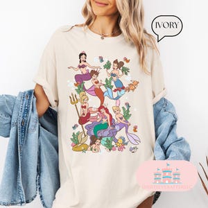 May include: Ivory-colored t-shirt featuring a colorful illustration of several mermaids in a whimsical underwater scene. The mermaids have various hair colors and wear seashell tops and mermaid tails. The shirt has the text "IVORY" in a speech bubble.