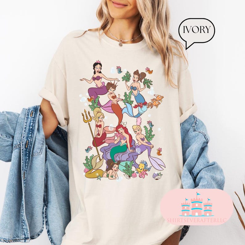 May include: Ivory-colored t-shirt featuring a colorful illustration of several mermaids in a whimsical underwater scene. The mermaids have various hair colors and wear seashell tops and mermaid tails. The shirt has the text "IVORY" in a speech bubble.