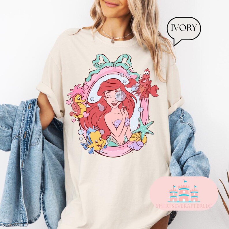 K&ouml;nnte beinhalten: Elfenbeinfarbenes T-Shirt mit einer farbenfrohen Grafik einer rothaarigen Meerjungfrau und Meeresbewohnern in einem rosa und blauen Rahmen. Das Shirt hat hochgekrempelte &Auml;rmel und das Wort "IVORY" steht in einer Sprechblase.