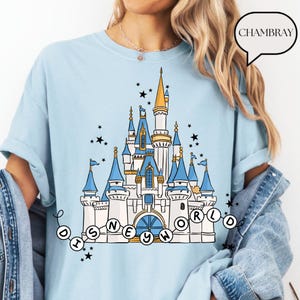 May include: Light blue t-shirt featuring a detailed illustration of a fairytale castle with blue turrets, gold accents, and the text "DISNEY WORLD" in a circular design. Black star accents surround the castle. The shirt has rolled-up sleeves.