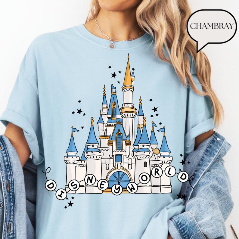 May include: Light blue t-shirt featuring a detailed illustration of a fairytale castle with blue turrets, gold accents, and the text "DISNEY WORLD" in a circular design. Black star accents surround the castle. The shirt has rolled-up sleeves.