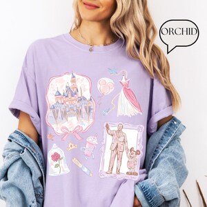 Op de afbeelding: Een lichtpaars T-shirt met verschillende illustraties van een kasteel, jurk, ballonnen en personages. Het woord "ORCHID" staat in een tekstballon. De illustraties zijn in pastelkleuren, wat zorgt voor een grillig en speels ontwerp.