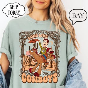 May include: A light green t-shirt featuring a cartoon cowboy riding a horse with the words "Long Live Cowboys". The design includes a cowboy character and a horse in a desert landscape.
