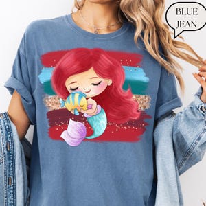 Puede incluir: Camiseta azul vaquera con una ilustraci&oacute;n de sirena de dibujos animados. La sirena tiene el pelo rojo y una cola turquesa, abrazando a un pez amarillo y azul. El dise&ntilde;o incluye pinceladas rojas, turquesas y doradas. El texto "BLUE JEAN" est&aacute; en una burbuja.