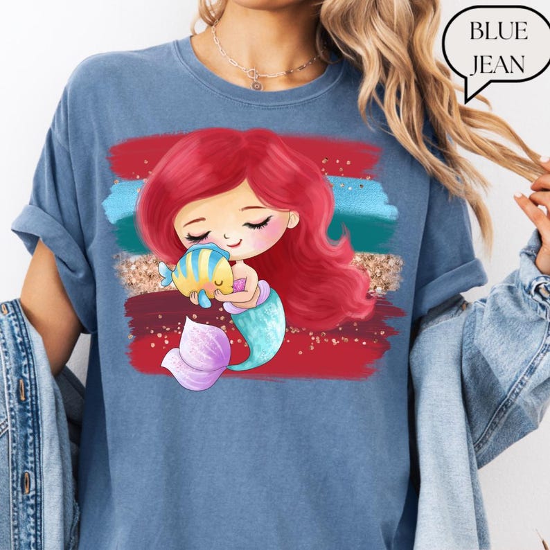 Puede incluir: Camiseta azul vaquera con una ilustraci&oacute;n de sirena de dibujos animados. La sirena tiene el pelo rojo y una cola turquesa, abrazando a un pez amarillo y azul. El dise&ntilde;o incluye pinceladas rojas, turquesas y doradas. El texto "BLUE JEAN" est&aacute; en una burbuja.