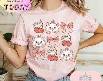 May include: Pink t-shirt with a repeated pattern of a cartoon cat with a pink bow, cherries, and green leaves. The shirt has a crew neck and short sleeves.