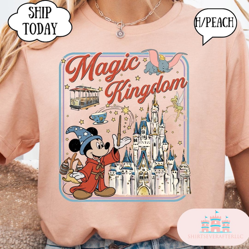 May include: A light peach-colored t-shirt featuring a graphic of Mickey Mouse as a sorcerer, a castle, and the words "Magic Kingdom." The design includes Dumbo, Tinkerbell, and a cable car. The shirt has a round neck.