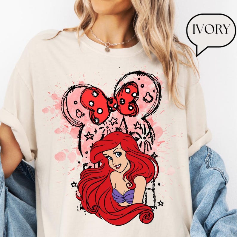 May include: Ivory-colored t-shirt featuring a graphic of Ariel from The Little Mermaid. The design includes a red bow with white polka dots, a pink watercolor splash, and black outline of Mickey Mouse ears. The word "IVORY" is in a speech bubble.