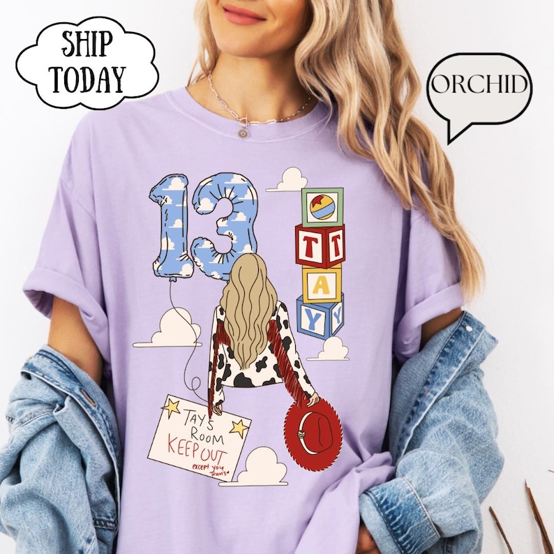 May include: A light purple t-shirt featuring a graphic of a person with long blonde hair, a cow-print jacket, and a red hat. The design includes a blue cloud-patterned number 13 balloon, stacked alphabet blocks, and a sign reading Tays Room KEEP OUT.