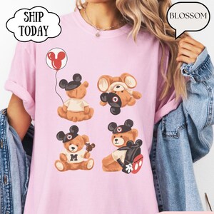 May include: A light pink t-shirt featuring a graphic print of four brown teddy bears wearing black mouse ear hats. One bear holds a red balloon, another wears a white shirt with an M, and one carries a black backpack with red accents.