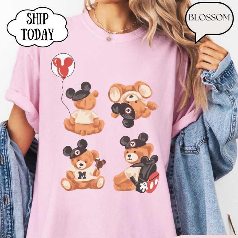 May include: A light pink t-shirt featuring a graphic print of four brown teddy bears wearing black mouse ear hats. One bear holds a red balloon, another wears a white shirt with an M, and one carries a black backpack with red accents.
