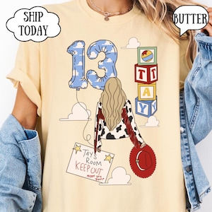 May include: A butter yellow t-shirt featuring a graphic of a person with long blonde hair, a cow-print jacket with red fringe, and a red hat. The design includes a blue cloud-patterned number 13 balloon, alphabet blocks, and a sign reading TAY'S ROOM KEEP OUT except you Travis.