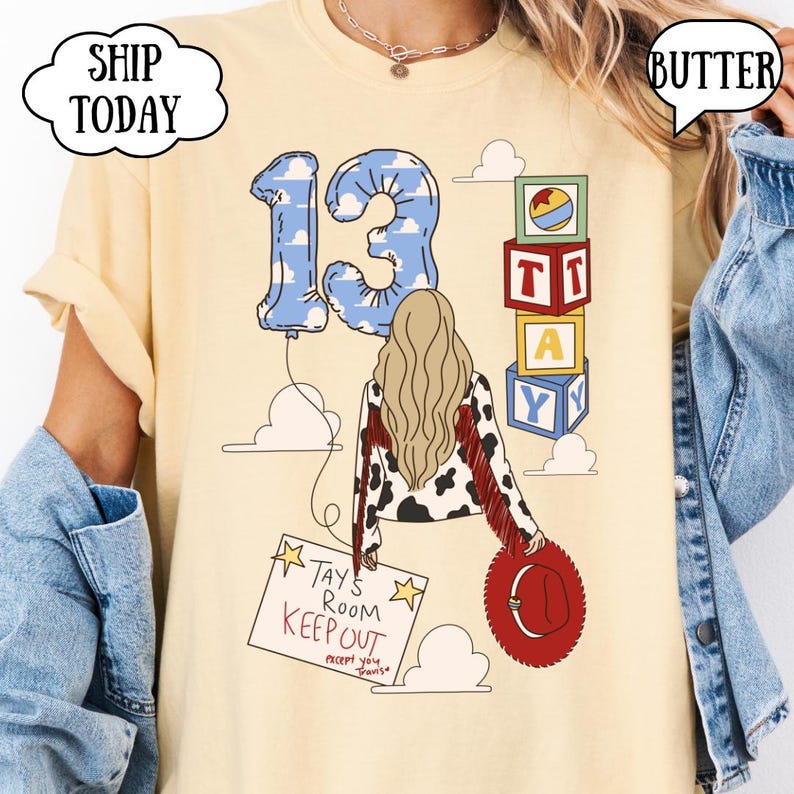 May include: A butter yellow t-shirt featuring a graphic of a person with long blonde hair, a cow-print jacket with red fringe, and a red hat. The design includes a blue cloud-patterned number 13 balloon, alphabet blocks, and a sign reading TAY'S ROOM KEEP OUT except you Travis.