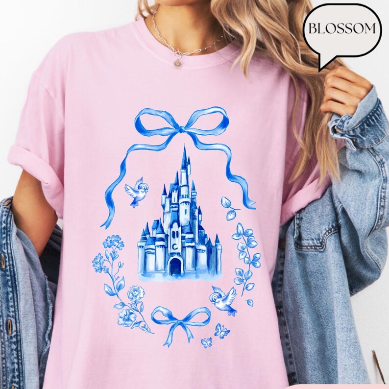 以下が含まれることがあります： 水彩画の青いお城のデザインが特徴の薄いピンク色のTシャツ。花のリース、リボン、鳥に囲まれています。「BLOSSOM」という言葉が吹き出しの中にあります。シャツは薄いピンク色です。