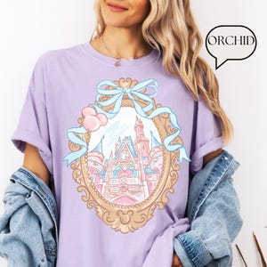 Puede incluir: Una camiseta de color lila con una ilustraci&oacute;n detallada de un castillo de cuento de hadas dentro de un marco decorativo, adornada con un lazo azul claro y detalles en rosa. La palabra "ORCHID" est&aacute; en una burbuja. La camiseta tiene mangas enrolladas.