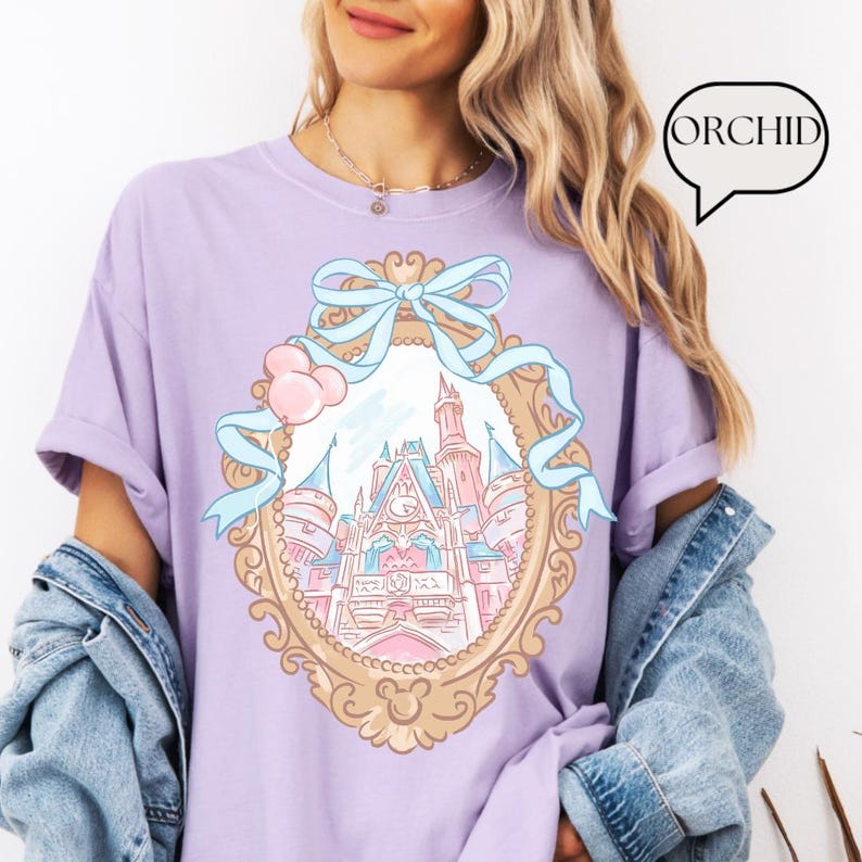 Puede incluir: Una camiseta de color lila con una ilustraci&oacute;n detallada de un castillo de cuento de hadas dentro de un marco decorativo, adornada con un lazo azul claro y detalles en rosa. La palabra "ORCHID" est&aacute; en una burbuja. La camiseta tiene mangas enrolladas.