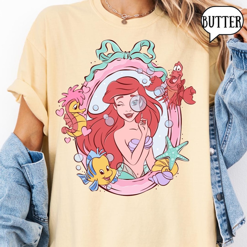 May include: A light yellow t-shirt featuring a cartoon illustration of Ariel from The Little Mermaid. The design includes Ariel, Flounder, Sebastian, a seahorse, and a starfish, all within a pink and teal frame. The word "BUTTER" is in a speech bubble.