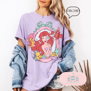 K&ouml;nnte beinhalten: Ein lilafarbenes T-Shirt mit einer Cartoon-Illustration einer rothaarigen Meerjungfrau mit einer Lupe und Meeresbewohnern. Das Design ist von einem rosa-gr&uuml;nen Rahmen mit einer Schleife umgeben. Das Wort "ORCHID" befindet sich in einer Sprechblase.