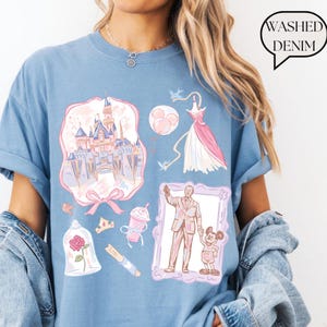 Op de afbeelding: Lichtblauw T-shirt met een afbeelding van een kasteel, jurk en andere illustraties in roze en paars. Het shirt heeft de tekst "Washed Denim" in een tekstballon.