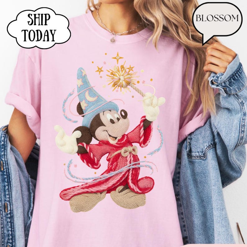 May include: A pale pink t-shirt with a cartoon Mickey Mouse in a wizard outfit. Mickey is wearing a blue hat with a crescent moon, a red robe, and holding a wand with a starburst design. The text "BLOSSOM" is in a speech bubble.