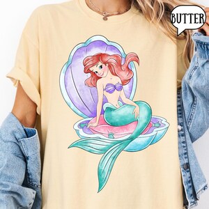 May include: A pale yellow t-shirt with a cartoon mermaid illustration. The mermaid has red hair, a purple seashell bra, and a teal tail, and is seated in a seashell. The word "BUTTER" is in a speech bubble.